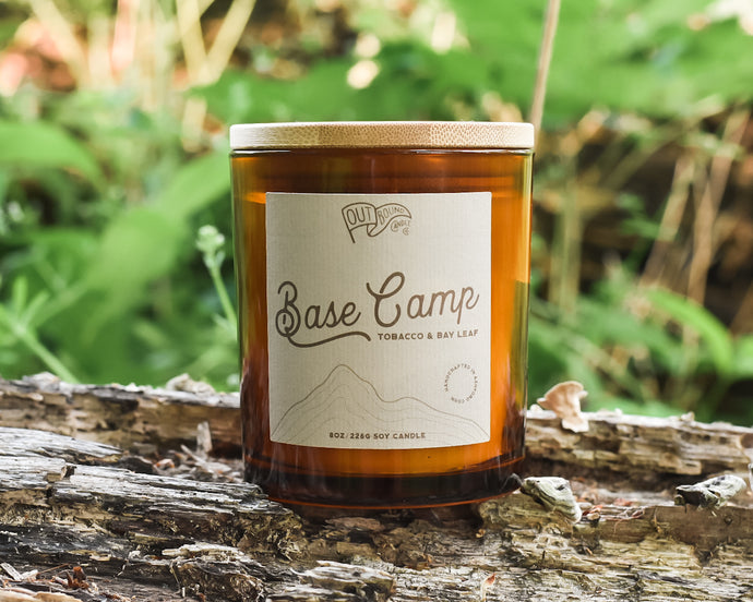A candle called Base camp with notes of tobacoo and bay leaf in an amber jar and a bamboo lid sits on a log in the lush green woods