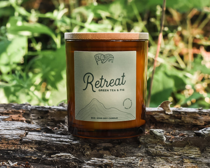 A candle called Retreat with notes of Green Tea and Fig in an amber jar and a bamboo lid sits on a log in the lush green woods