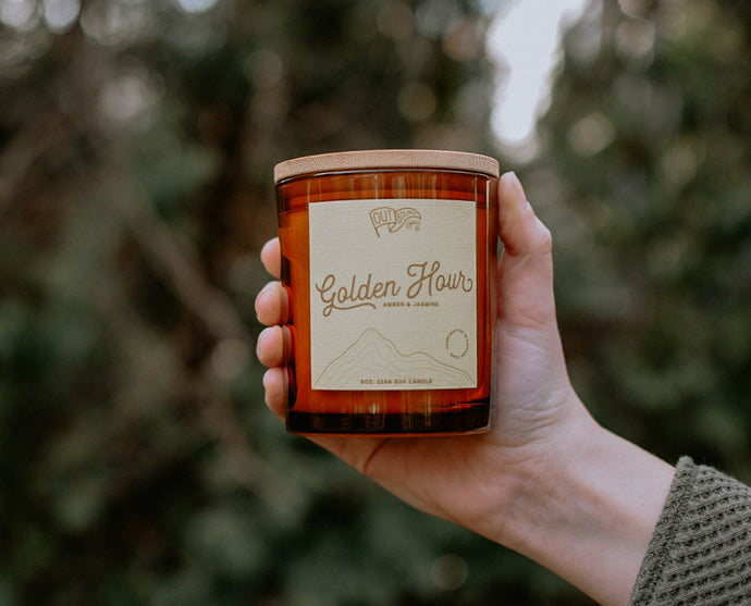 A candle called Golden hour with notes of Jasmine and Amber in an amber jar with a bamboo lid is held out in the green tree line glowing in the afternoon sun