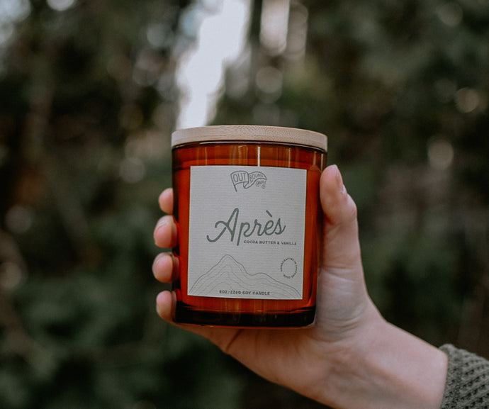 A candle called Après with notes of Coco Butter and Vanilla in an amber jar and bamboo lid is held out in the green tree line 