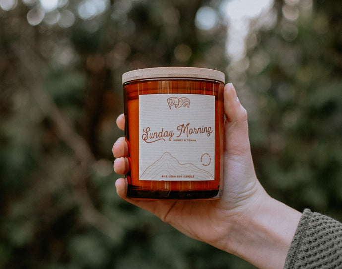 A candle called Sunday Morning with notes of Spiced Honey and Tonka in an amber jar and bamboo lid is held out in the green tree line 