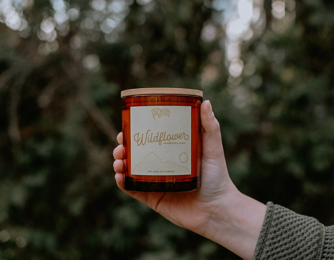 A candle called Wildflower with notes of Sweetgrass and Dandelion in an amber jar and bamboo lid is held out in the green tree line 